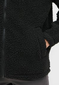 Veste en polaire noire avec un tissu texturé, fermeture éclair avant et poches latérales. Le design comprend une coupe décontractée et des poignets côtelés.