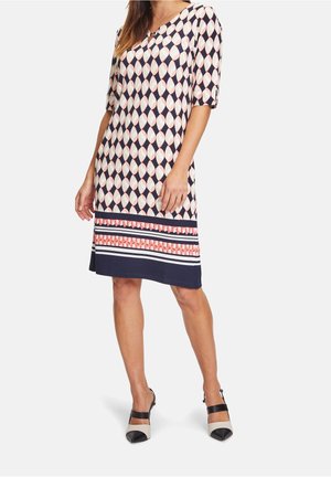 Kobieta ubrana w sukienkę do kolan z granatowo-biało-koralowym geometrycznym wzorem i rękawami do łokcia, zestawioną z czarno-białymi butami na szpic.