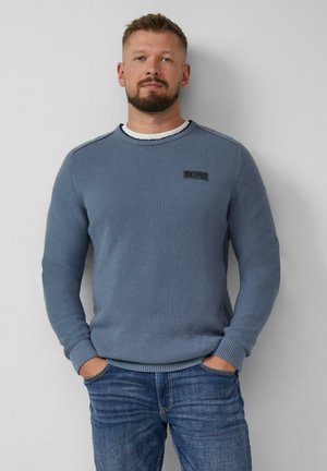 Man met kort haar en baard, draagt een blauwe gebreide trui over een wit shirt en blauwe jeans, staat met zijn handen in zijn zakken tegen een grijze muur.