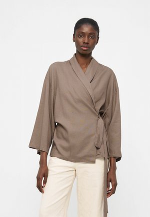 Moteris, dėvinti taupe spalvos permatomos stiliaus palaidinę su plačiomis rankovėmis ir kreminius aukšto liemens kelnes, stovinti priešais paprastą šviesią foną.