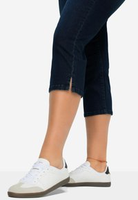 Sneaker bianchi con tomaia in pelle liscia, accenti in suede grigio e suola in gomma marrone. Indossati con pantaloni in denim blu scuro a metà polpaccio.