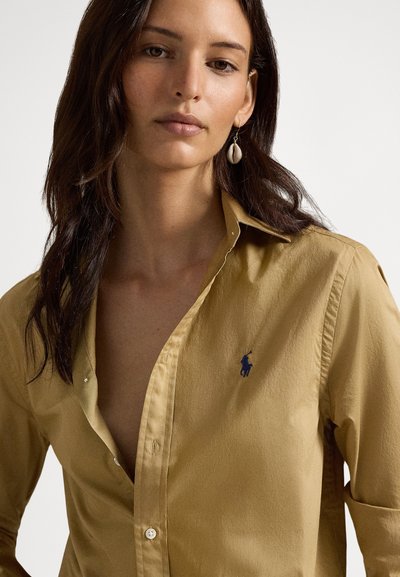 Žena s dlhými tmavými vlasmi, oblečená v béžovej košeli na gombíky s námornícky modrým vyšívaným logom a náušnicami v tvare mušle.