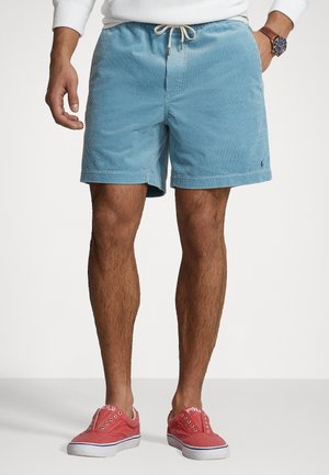 Homme portant un short en velours côtelé bleu clair avec cordon de serrage et des baskets rouges sans lacets, debout devant un fond blanc uni.