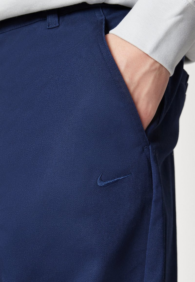 Pantaloni blu navy realizzati in un tessuto liscio con un design elegante. Presentano un logo Nike blu ben visibile e tasche laterali.