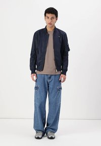 Marineblå bomberjakke med glidelås, ribbede mansjetter og en sidelomme, kombinert med løse blå cargo-jeans og grå sneakers.
