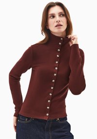 Ribbet burgunderfarvet sweater med høj krave, der har en række metallic knapper langs venstre side og lange ærmer.