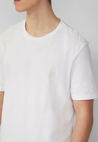 Weißes Baumwoll-T-Shirt mit einem kleinen gelben runden Logo auf der linken Brust. Kurze Ärmel und ein Rundhalsausschnitt. Glatte Textur.