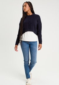 Granatowy sweter z dzianiny w warkocze noszony na białej koszulce, sparowany z jasnoniebieskimi, dopasowanymi dżinsami i beżowymi sneakerm, z teksturowanym wzorem.