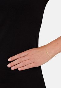 Guldarmband med en delikat kedjedesign, som har små runda dekorationer. Bärs på en handled mot en svart skjortbakgrund.