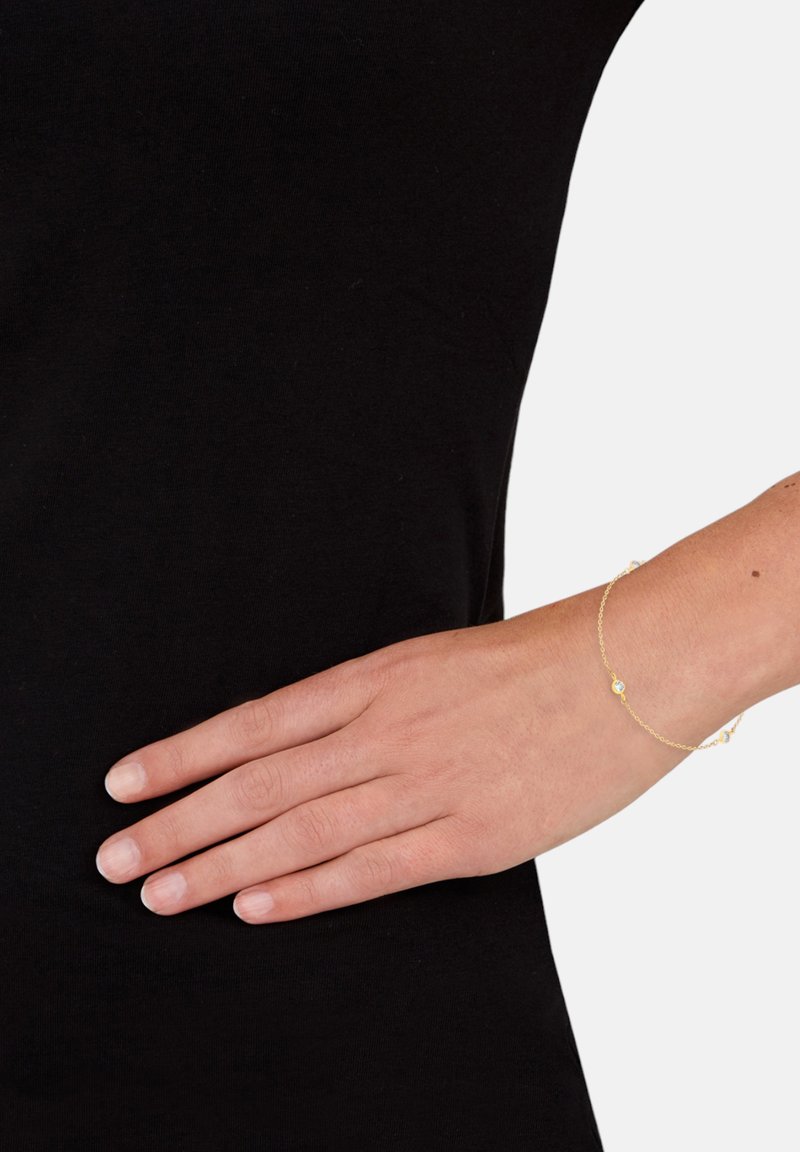 Guldarmband med en delikat kedjedesign, som har små runda dekorationer. Bärs på en handled mot en svart skjortbakgrund.