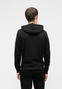 Fekete kapucnis pulóver lekerekített kapucnival, bordás mandzsettákkal és szegéllyel. Sima textúra; nincs látható minta vagy díszítés. A modell hátulról látható.