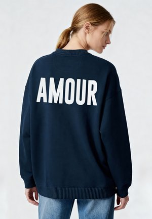 Vrouw met lichtbruin haar in een paardenstaart draagt een donkerblauwe sweatshirt met "AMOUR" in grote witte letters op de rug en lichtblauwe spijkerbroek.
