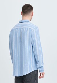 Chemise bleu clair à boutons avec rayures blanches verticales, manches longues, col pointu, et ourlet légèrement arrondi. Confectionnée en tissu doux.