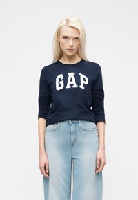 Νέα γυναίκα με ίσια ξανθά μαλλιά που φοράει μπλε σκούρο φούτερ GAP και ανοιχτόχρωμο τζιν υψηλής μέσης, στέκεται μπροστά σε απλό φόντο.
