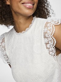 Prim-plan al unei persoane zâmbitoare care poartă un top alb din dantelă cu modele florale și mâneci și decolteu din dantelă cu margini ondulate.