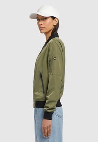 Giacca bomber verde oliva con colletto e polsini neri, tasche con zip e tessuto dalla texture elegante. Abbinata a jeans azzurri chiari e un cappellino bianco.