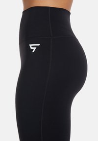 High-waisted black leggings made of stretchy fabric with a smooth texture. Features a white logo on the left side and decorative seam lines.