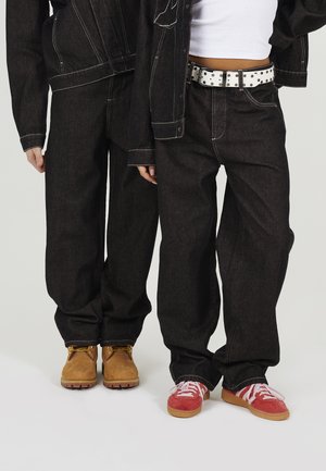 Dwie osoby w ciemnych jeansach; jedna w brązowych botkach, druga w czerwonych trampkach i białym krótkim topie z paskiem w groszki.