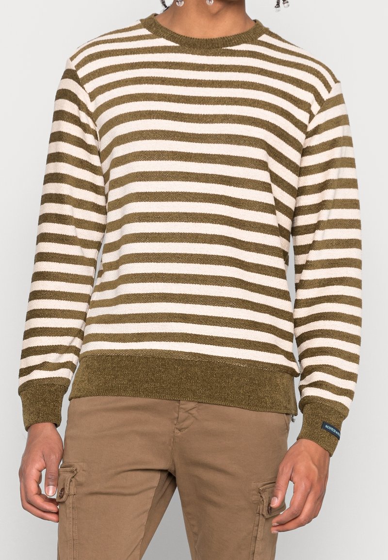 Homme portant un pull rayé horizontalement olive et blanc et un pantalon cargo marron clair, debout devant un fond uni.