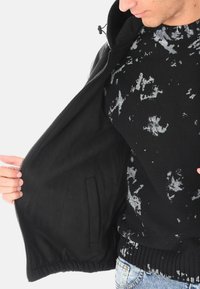 Persona che indossa un maglione a strisce nere e grigie e jeans blu, mentre tiene aperta una giacca nera per mostrare la sua tasca interna.