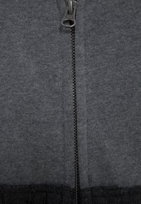 Primer plano de un zip de tela gris oscuro con dientes de metal entrelazados visibles y una pestaña de tirador de YKK.