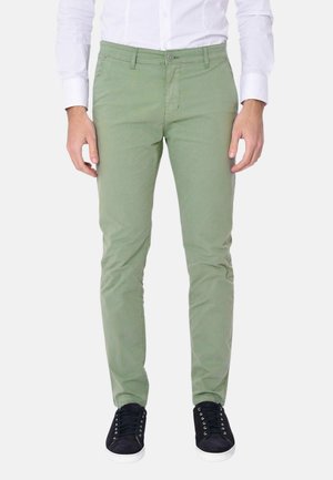 Uomo che indossa pantaloni verde chiaro aderenti, camicia bianca a maniche lunghe e sneakers nere con suola bianca, in piedi contro uno sfondo semplice.