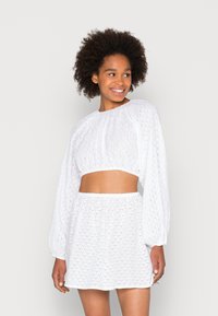 Blusa branca curta com mangas bufantes e padrão de ilhós, combinada com uma saia branca correspondente com cintura elástica e design em camadas.