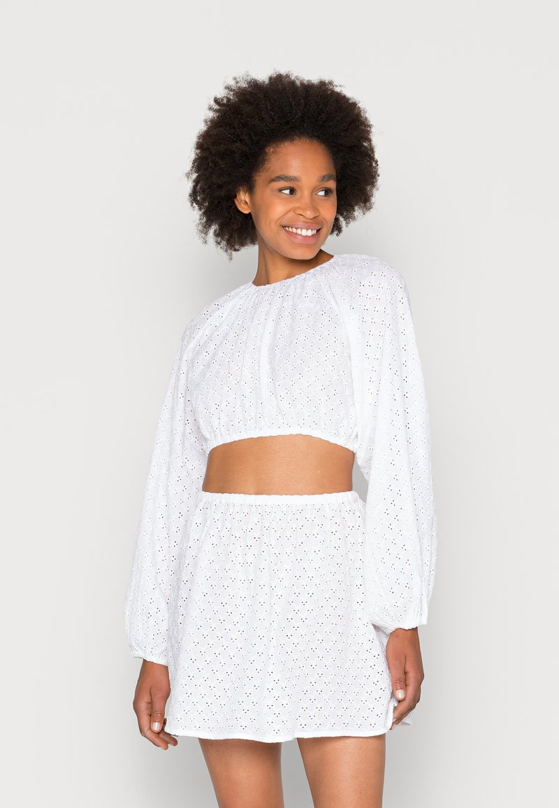 Blusa branca curta com mangas bufantes e padrão de ilhós, combinada com uma saia branca correspondente com cintura elástica e design em camadas.