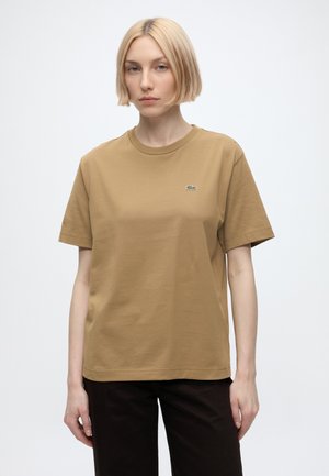 Femme aux cheveux blonds courts portant un t-shirt marron uni avec un petit logo crocodile sur la poitrine et un pantalon noir sur fond blanc.