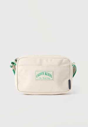 Sac bandoulière en tissu beige avec un patch logo vert, une fermeture éclair et une sangle accentuée verte. Design rectangulaire compact.