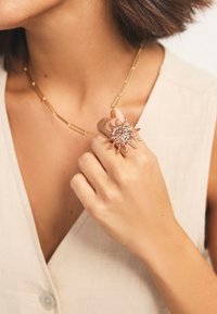 Gold chain necklace with elongated links. Sun-shaped ring featuring intricate details and shimmering accents on a hand. Neutral fabric background.