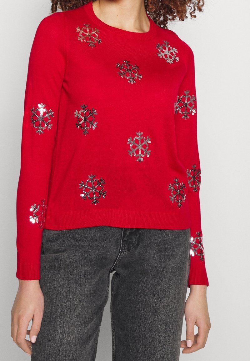 Pull rouge à manches longues avec des motifs de flocons de neige en sequins argentés, porté avec un jean gris foncé.