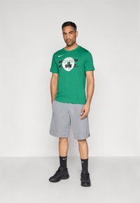 Πράσινο βαμβακερό μπλουζάκι με το λογότυπο "Celtics", συνδυασμένο με γκρι σορτς. Μαύρα αθλητικά παπούτσια και κάλτσες με το λογότυπο της Nike ολοκληρώνουν το ντύσιμο.