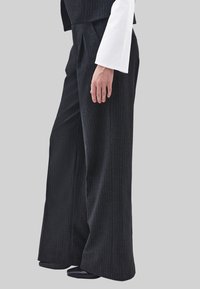 Zwarte wijd uitlopende broek met krijtstreep, vervaardigd uit een gestructureerde stof, voorzien van plooien en gecombineerd met een contrasterend wit topje en zwarte schoenen.