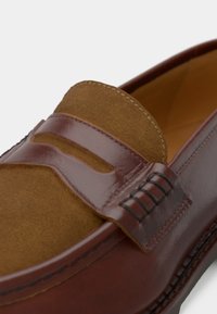 Mocassim castanho em pele e camurça com design de bico em forma de bico e detalhe de tira na parte superior, mostrando um close-up das costuras e texturas.