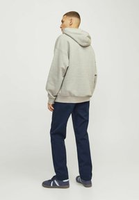 Mand iført en grå hættetrøje, mørkeblå bukser og mørkeblå sneakers, stående med ryggen vendt mod en ensfarvet lys baggrund.