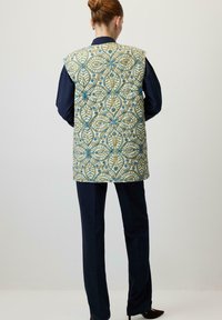 Gepatterneerde vest in groen, geel en blauw met bladmotieven; heeft een mouwloze ontwerp en knoopsluitingen op de achterkant. Gecombineerd met marineblauwe broek.