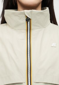 Κοντινό πλάνο ενός μπεζ σακακιού με ψηλό γιακά, κάθετο φερμουάρ με ρίγες σε πορτοκαλί, κίτρινο, ναυτικό μπλε και λευκό, και ένα μικρό λογότυπο στο στήθος.