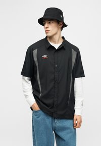 Νεαρός άνδρας με μαύρο καπέλο τύπου κάλπη, μαύρο πουκάμισο με κοντά μανίκια και γκρι ρίγες, λευκό πουκάμισο με μακριά μανίκια από κάτω και μπλε τζιν.