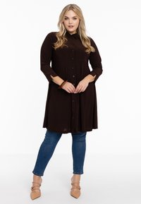 Braunes Hemdkleid mit Knöpfen, langen Ärmeln, A-Linien-Form und seitlichen Schlitzen, kombiniert mit blauen Skinny-Jeans und nudefarbenen spitzen Ballerinas.