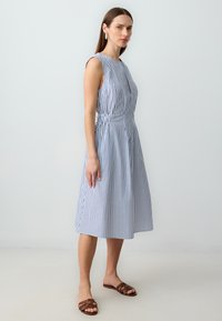 Blau-weiß gestreiftes ärmelloses Kleid mit taillierter Passform und weitem Rock, aus leichtem Stoff gefertigt. Braune flache Sandalen runden den Look ab.