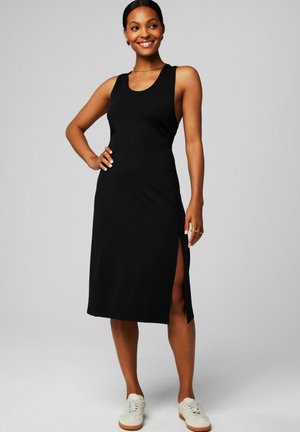 Smiling woman wearing a sleeveless black midi dress with side slit and white sneakers, standing with one hand on hip against gray background.