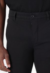 Black cotton trousers featuring a front button closure, side pockets, and a slim fit design. Textured fabric with a smooth finish.