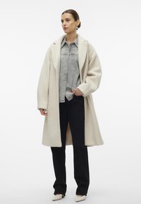 Crème-kleurige lange jas van zachte, gestructureerde stof met een brede kraag, gelaagd over een grijze denimshirt en donkere broek. Puntige beige schoenen.