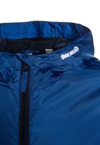 Niebieska lekka kurtka o gładkim wykończeniu, z ciemnym zamkiem błyskawicznym, regulowanym kapturem ze sznurkiem oraz widoczną siateczkową podszewką wewnątrz.