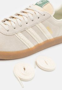 Béžové semišové tenisky s třemi pletenými bílými pruhy, gumovou podrážkou a světle krémovými tkaničkami. Na boku zlaté logo "Gazelle".