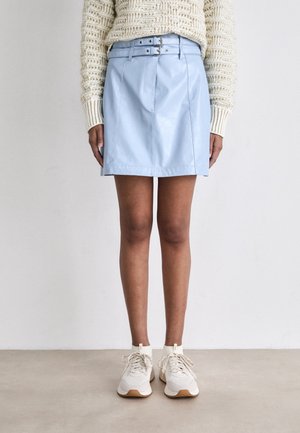 Personne portant un pull en maille crème, une mini-jupe en cuir bleu clair ceinturée et des baskets blanches, debout contre un mur blanc uni.