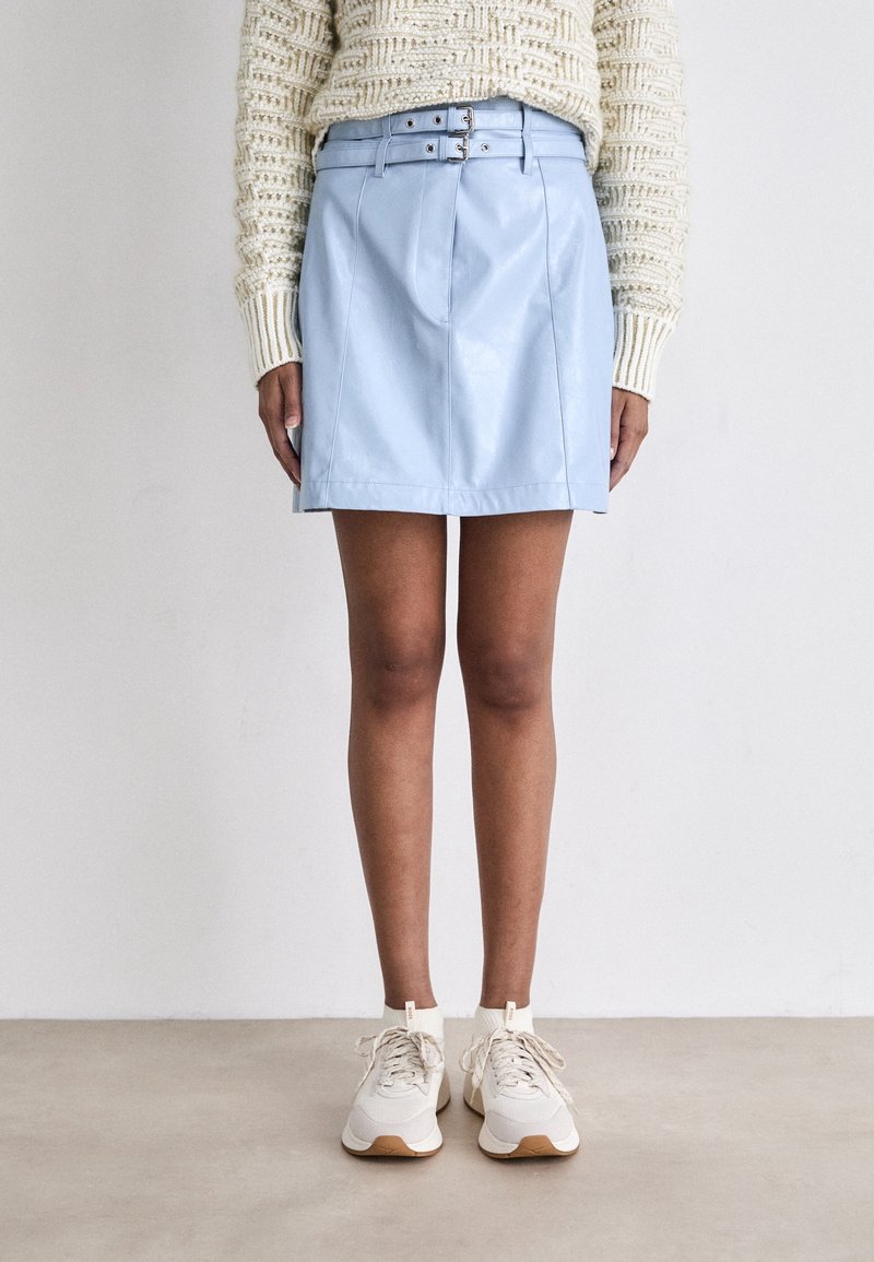 Personne portant un pull en maille crème, une mini-jupe en cuir bleu clair ceinturée et des baskets blanches, debout contre un mur blanc uni.