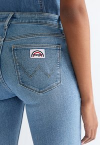 Detail zadní části osoby nosící světle modré džíny Wrangler s viditelnou značkovou nášivkou na pravé zadní kapse.