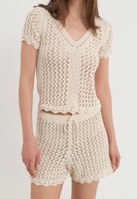 Dwu-częściowy zestaw dzianinowy w kolorze ecru, z karbowanym dekoltem i krótkimi rękawami. Wzorzysty materiał o luźnym kroju oraz szorty ze ściągaczem.